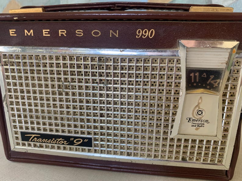 May include: Vintage Emerson 990 transistor radio in a brown leatherette case. The radio features a silver speaker grill, a tuning dial, and the text "Transistor '9'". The Emerson logo is visible on the dial.