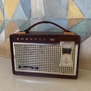 May include: A vintage Emerson 990 transistor radio in a brown leatherette case. The radio has a silver speaker grill, a tuning dial, and a carrying handle. The text "Emerson" and "Transistor '9'" are visible.