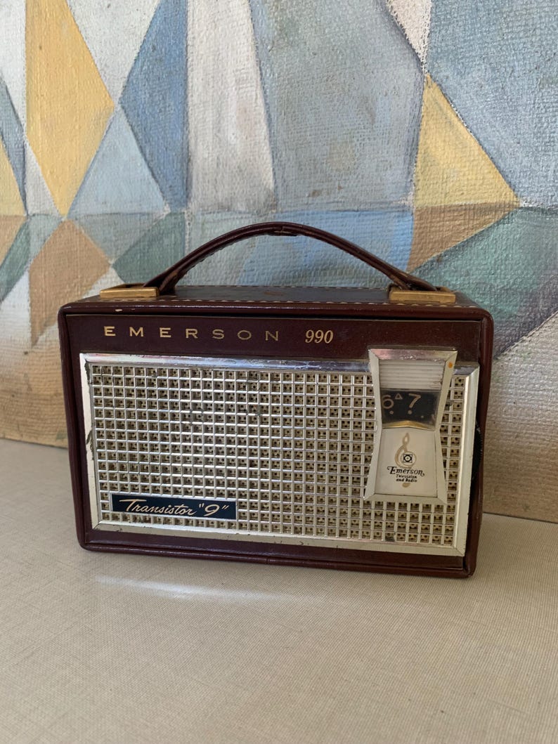May include: A vintage Emerson 990 transistor radio in a brown leatherette case. The radio has a silver speaker grill, a tuning dial, and a carrying handle. The text "Emerson" and "Transistor '9'" are visible.
