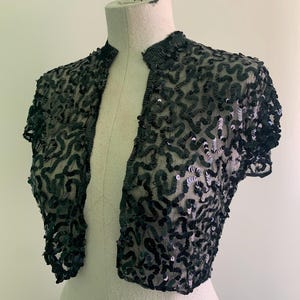 May include: A dark green sequined bolero jacket with short sleeves, displayed on a mannequin. The open-front jacket is covered in a pattern of sequins, creating a shimmering effect. The garment is designed to be worn over other clothing.