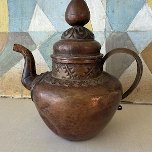 May include: A copper teapot featuring a rounded body, a curved spout, and a handle. The lid is topped with a finial and decorated with embossed leaf patterns around the neck and base of the lid, showcasing a weathered, dark metallic finish.