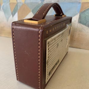 May include: A vintage brown leather Emerson radio with a handle and a speaker grill. The radio has a rectangular shape with a stitched design and gold-colored accents. The radio's face has the word "Emerson" printed on it.