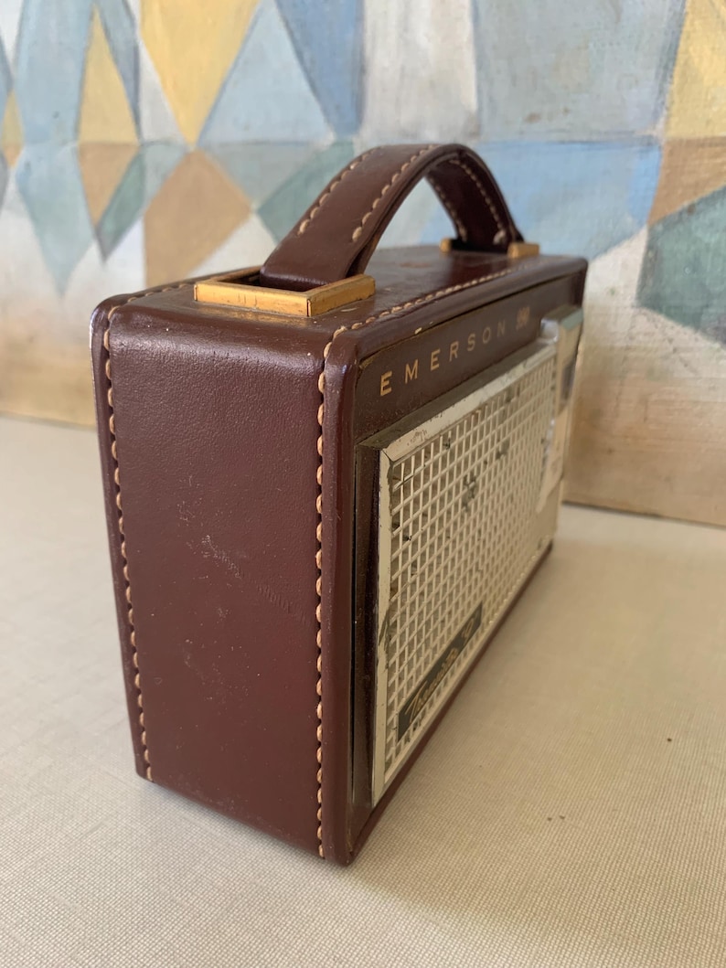May include: A vintage brown leather Emerson radio with a handle and a speaker grill. The radio has a rectangular shape with a stitched design and gold-colored accents. The radio's face has the word "Emerson" printed on it.