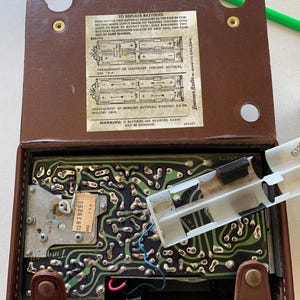 May include: Open brown vintage Emerson Radio with the back cover open, revealing the internal circuitry and battery compartment. The text on the inside cover provides instructions for replacing batteries. A white plastic battery holder is partially removed.