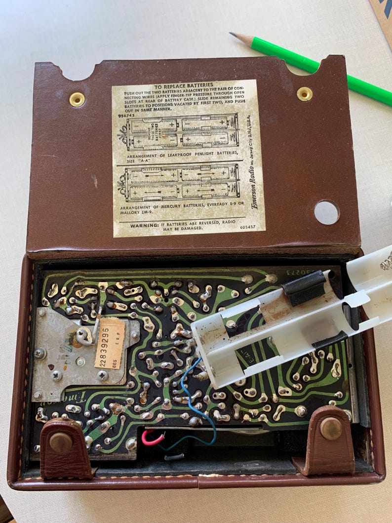 May include: Open brown vintage Emerson Radio with the back cover open, revealing the internal circuitry and battery compartment. The text on the inside cover provides instructions for replacing batteries. A white plastic battery holder is partially removed.