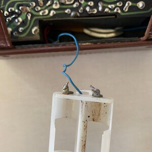 May include: A close-up of a white plastic battery holder with two metal terminals and a blue wire. The holder is dirty and shows signs of wear. The background includes a circuit board and a brown casing.