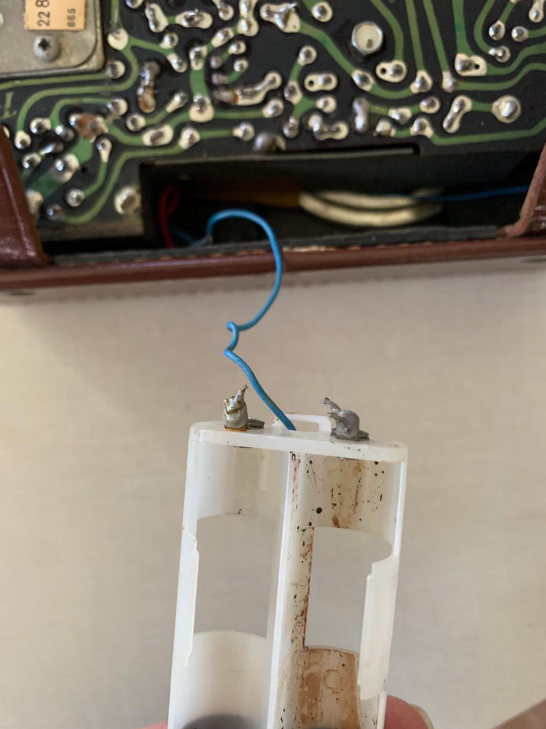 May include: A close-up of a white plastic battery holder with two metal terminals and a blue wire. The holder is dirty and shows signs of wear. The background includes a circuit board and a brown casing.