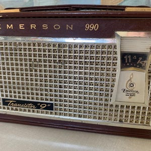 May include: Vintage Emerson 990 transistor radio in a brown leatherette case. The radio features a silver speaker grill, a tuning dial, and the text "Transistor 9". The Emerson logo is visible on the front.