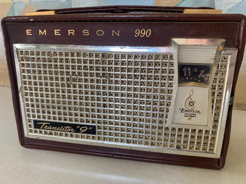May include: Vintage Emerson 990 transistor radio in a brown leatherette case. The radio features a silver speaker grill, a tuning dial, and the text "Transistor 9". The Emerson logo is visible on the front.