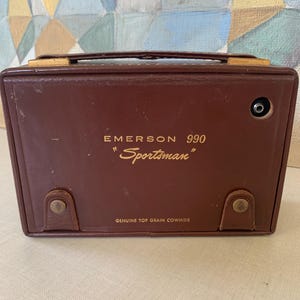 May include: A vintage Emerson 990 "Sportsman" radio in a brown leather case. The radio has a handle, metal accents, and the words "Genuine Top Grain Cowhide" are printed on the bottom.