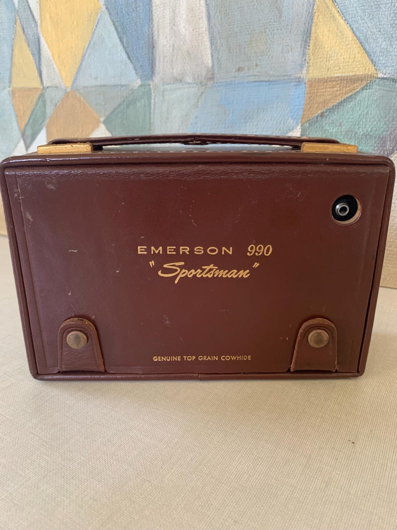 May include: A vintage Emerson 990 "Sportsman" radio in a brown leather case. The radio has a handle, metal accents, and the words "Genuine Top Grain Cowhide" are printed on the bottom.
