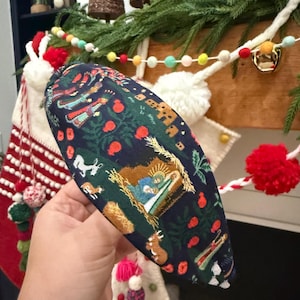 May include: A padded headband with a dark blue background and a Christmas-themed pattern. The pattern includes illustrations of a nativity scene, deer, and red berries. The headband is a festive accessory.