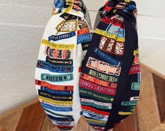 Op de afbeelding: Twee stoffen hoofdbanden met een boekprintontwerp. De ene hoofdband is wit met kleurrijke boektitels en de andere is zwart met kleurrijke boektitels.