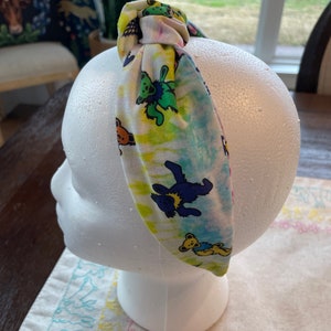 May include: A tie-dye headband with a colorful pattern of cartoon bears. The headband is tied in a knot on top of a white mannequin head.