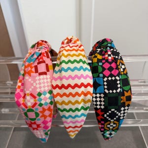 May include: Three knotted headbands featuring distinct fabric patterns. The left headband has a pink background with colorful geometric quilt-style shapes. The center headband displays horizontal wavy lines in orange, green, blue, and pink. The right headband has a black background with a grid of colorful geometric shapes.