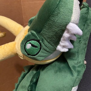 Op de afbeelding: Groene pluche draak met een gele hoorn en een witte bek. De draak heeft een groen oog met een zwarte pupil.