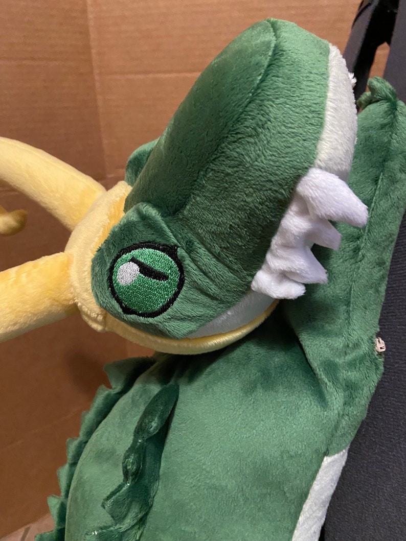Op de afbeelding: Groene pluche draak met een gele hoorn en een witte bek. De draak heeft een groen oog met een zwarte pupil.