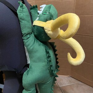 Op de afbeelding: Groene pluche dinosaurus rugzak met gele hoorns. De dinosaurus klimt op de rug van een zwarte rugzak.