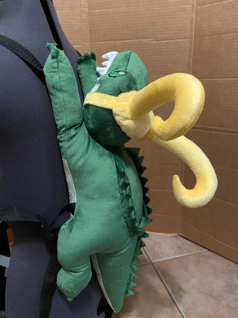 Op de afbeelding: Groene pluche dinosaurus rugzak met gele hoorns. De dinosaurus klimt op de rug van een zwarte rugzak.