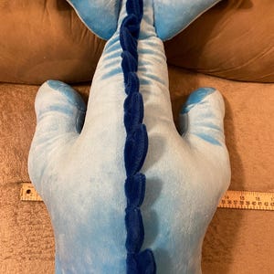 Puede incluir: Un juguete de peluche azul con forma de ballena con una aleta caudal azul y una aleta azul en la espalda. La ballena tiene un efecto degradado azul y blanco y una fila de p&uacute;as azules en la espalda.