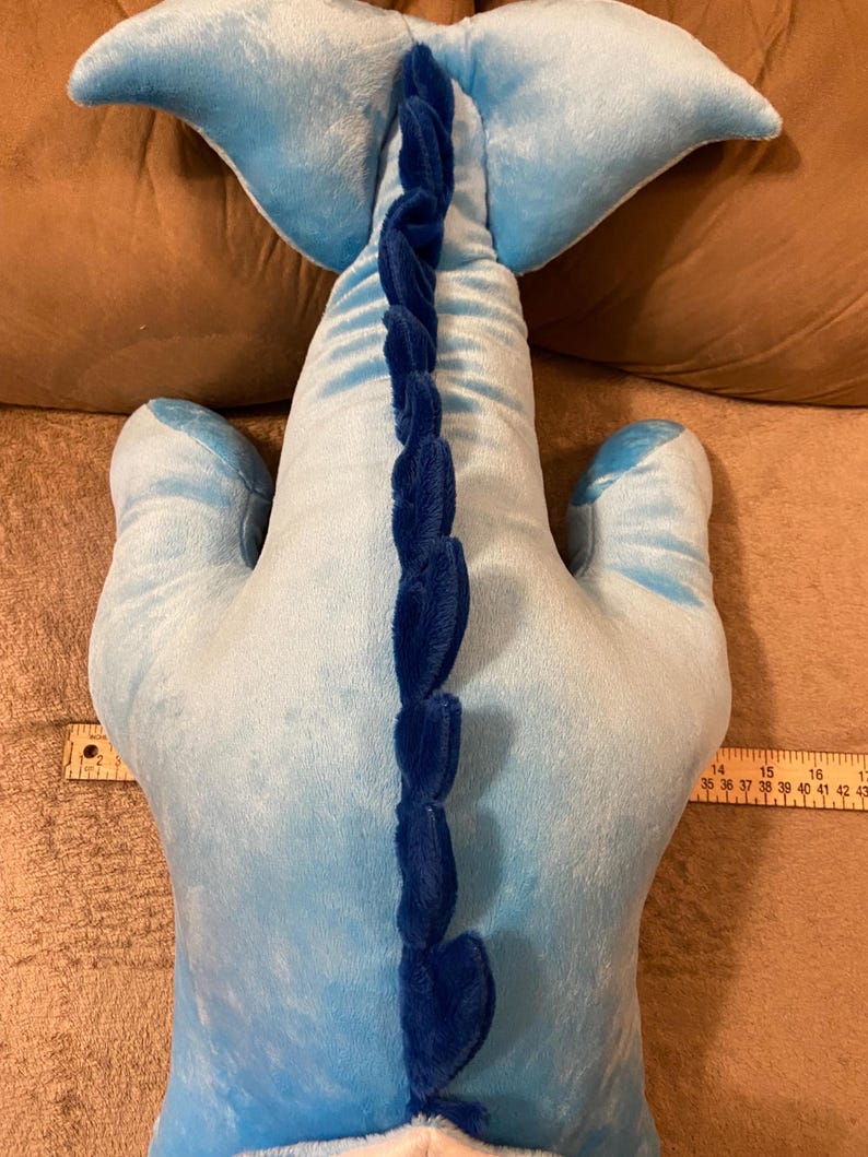 Puede incluir: Un juguete de peluche azul con forma de ballena con una aleta caudal azul y una aleta azul en la espalda. La ballena tiene un efecto degradado azul y blanco y una fila de p&uacute;as azules en la espalda.