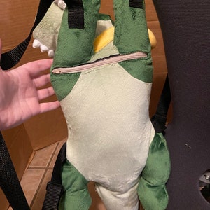 Op de afbeelding: Groene en gele pluche dinosaurus rugzak met zwarte banden. De rugzak heeft een rits aan de achterkant.