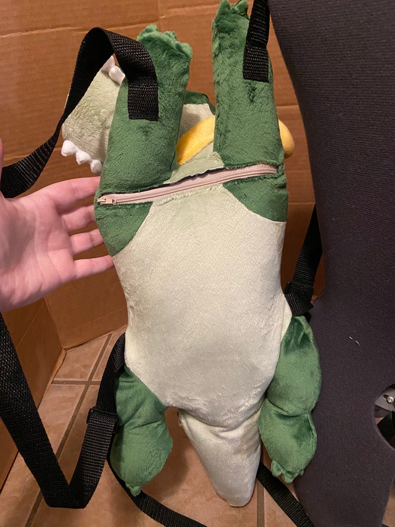Op de afbeelding: Groene en gele pluche dinosaurus rugzak met zwarte banden. De rugzak heeft een rits aan de achterkant.