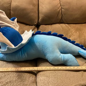 Puede incluir: Un juguete de peluche azul y blanco con forma de drag&oacute;n, acostado en un sof&aacute; marr&oacute;n. El juguete mide aproximadamente 76 cm de largo.