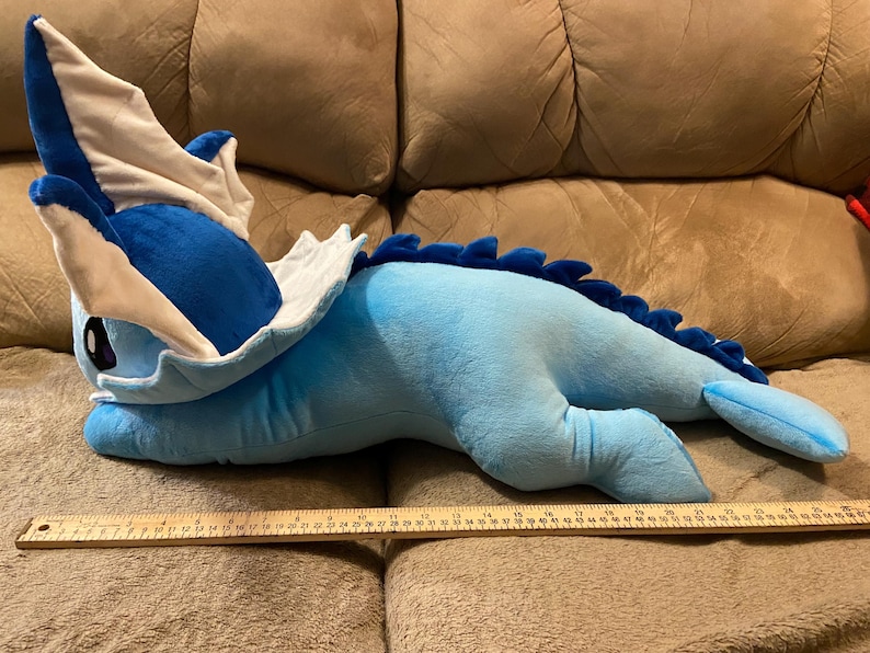 Puede incluir: Un juguete de peluche azul y blanco con forma de drag&oacute;n, acostado en un sof&aacute; marr&oacute;n. El juguete mide aproximadamente 76 cm de largo.
