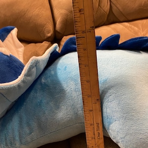 Puede incluir: Un juguete de peluche azul y blanco con forma de drag&oacute;n. El drag&oacute;n est&aacute; tumbado en un sof&aacute; marr&oacute;n. Una regla de madera est&aacute; al lado del drag&oacute;n, midiendo 28 cm de largo.
