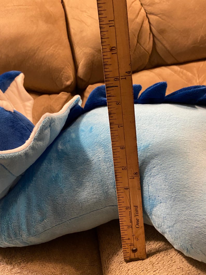 Puede incluir: Un juguete de peluche azul y blanco con forma de drag&oacute;n. El drag&oacute;n est&aacute; tumbado en un sof&aacute; marr&oacute;n. Una regla de madera est&aacute; al lado del drag&oacute;n, midiendo 28 cm de largo.