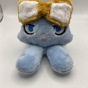 Puede incluir: Un peluche azul con forma de criatura con grandes ojos azules, una nariz rosa y una boca pequeña. Tiene un tocado amarillo y blanco. El juguete tiene dos brazos pequeños y dos piernas pequeñas.