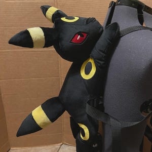 May include: A black Umbreon plush toy with yellow markings and red eyes, attached to a black backpack. The plush has yellow rings and accents on its ears and tail. The backpack has black straps.