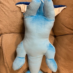 Puede incluir: Un juguete de peluche azul y blanco con forma de delf&iacute;n. El juguete est&aacute; tumbado de espaldas con las aletas y la cola extendidas.
