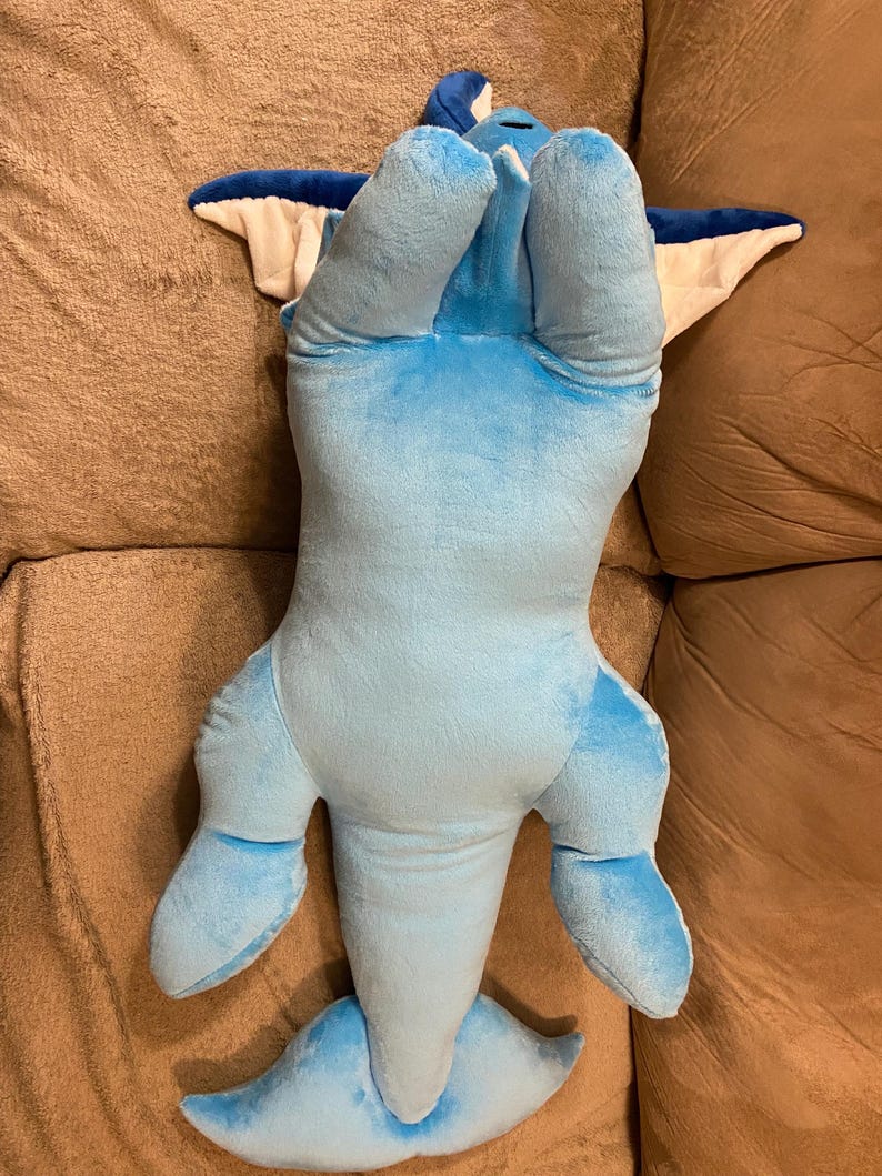 Puede incluir: Un juguete de peluche azul y blanco con forma de delf&iacute;n. El juguete est&aacute; tumbado de espaldas con las aletas y la cola extendidas.