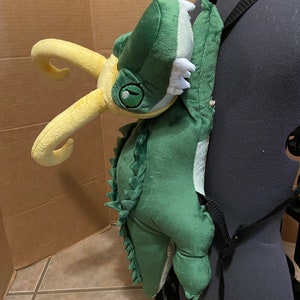 Op de afbeelding: Groene en gele pluche rugzak in de vorm van een dinosaurus met een tandenrij. De rugzak heeft verstelbare banden en een ritssluiting.