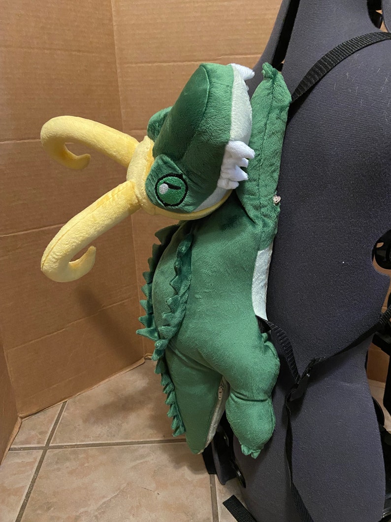 Op de afbeelding: Groene en gele pluche rugzak in de vorm van een dinosaurus met een tandenrij. De rugzak heeft verstelbare banden en een ritssluiting.