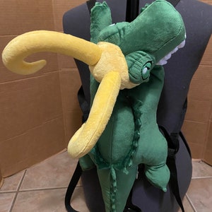 Op de afbeelding: Groene pluche alligator rugzak met een gele hoorn. De rugzak heeft verstelbare banden en is ontworpen om op de rug te worden gedragen.
