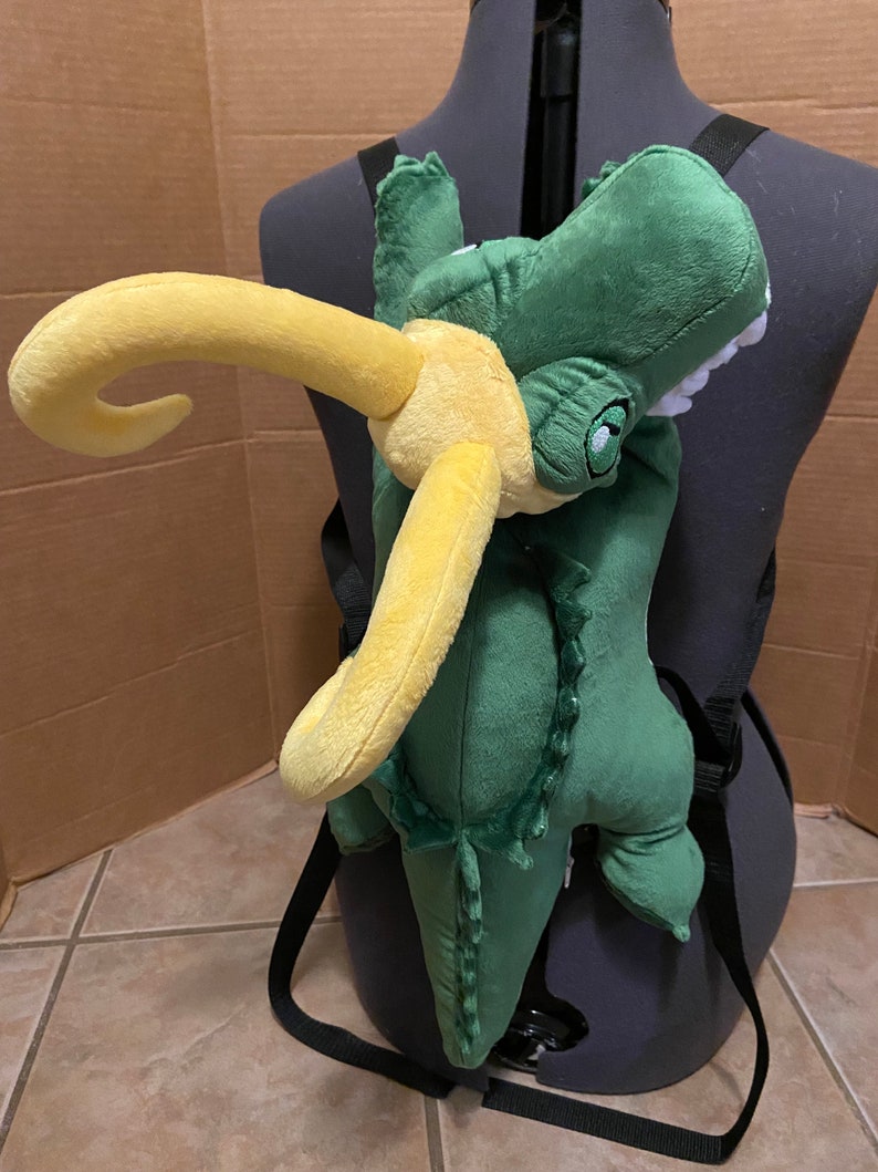 Op de afbeelding: Groene pluche alligator rugzak met een gele hoorn. De rugzak heeft verstelbare banden en is ontworpen om op de rug te worden gedragen.