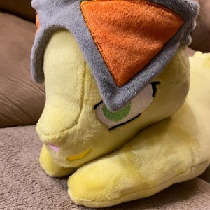May include: A plush yellow cat toy with a gray and orange head covering. The cat has green eyes and a pink nose. The toy is made of soft material and is resting on a brown surface.