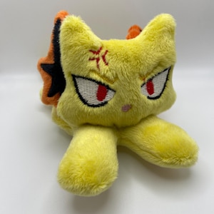 May include: A yellow plush toy cat with an angry expression. The cat has red eyes, a pink nose, and a red symbol on its forehead. The ears are yellow with orange and black accents. The toy is lying down.