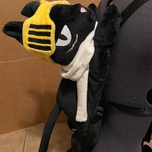 Może przedstawiać: Czarny pluszowy plecak w kształcie kota z żółtym i czarnym hełmem i białym szalikiem. Plecak ma regulowane paski.