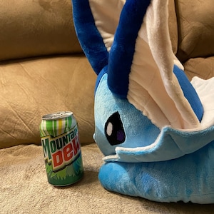 Puede incluir: Un juguete de peluche azul y blanco con orejas grandes, que se asemeja a un personaje de dibujos animados, est&aacute; acostado en un sof&aacute; marr&oacute;n junto a una lata de Mountain Dew.
