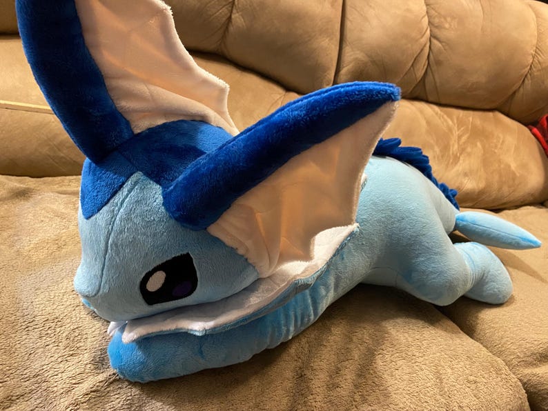 Puede incluir: Un juguete de peluche azul y blanco que se asemeja a una criatura m&iacute;tica con orejas grandes y una cola larga. El juguete est&aacute; acostado en un sof&aacute; marr&oacute;n.