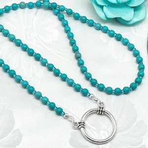 May include: A necklace featuring a long strand of round turquoise beads interspersed with smaller translucent blue glass beads. The necklace includes a large, circular silver-toned metal ring pendant attached to the beaded strand with silver findings.