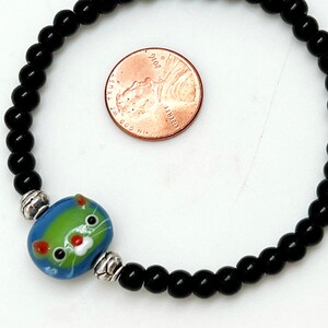 May include: A black beaded bracelet with a silver charm featuring a blue, green, and white cat face. The bracelet is next to a US penny for size reference.