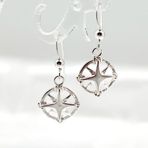 May include: Silver dangle earrings with a compass design. Each earring features a circular pendant with a star in the center and a decorative border.