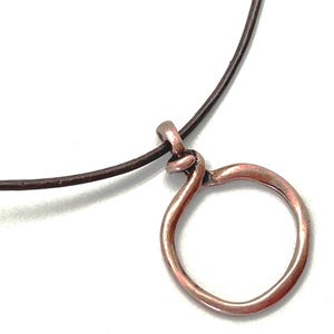 May include: A dark brown leather cord necklace featuring a circular copper pendant. The pendant has an irregular, hand-formed shape with a metallic, reddish-brown finish and is attached to the cord with a simple loop at the top of the circle.