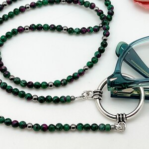 May include: A green and purple gemstone beaded eyeglass chain with a silver ring and clasp. The chain is made of small round beads and is approximately 30 inches long.