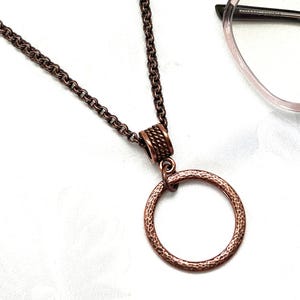 May include: A copper-colored necklace with a textured circular pendant. The chain is made of small, linked rings. The pendant has a hammered texture. The necklace is displayed on a white surface, with a pair of eyeglasses in the background.